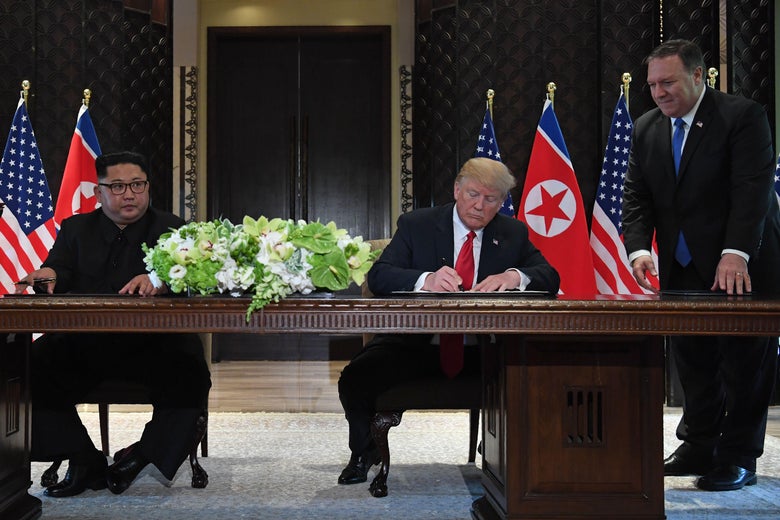 Kim Jong-un and Donald Trump sit at a table and sign the joint statement, with Mike Pompeo standing over Trump.